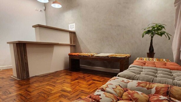 A modern hotel room with a wooden herringbone floor, a sofa with a leaf pattern, a small table, a potted plant, and a reception desk. - Morada City Hostel (São Paulo)