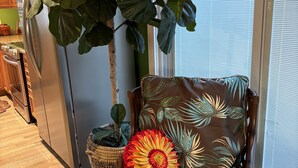 A wooden chair with a cushion and a decorative pillow, a large potted plant, and a kitchen area with wooden cabinets and a refrigerator. - Renovated Fisherman’s Cottage  (Lower Township)