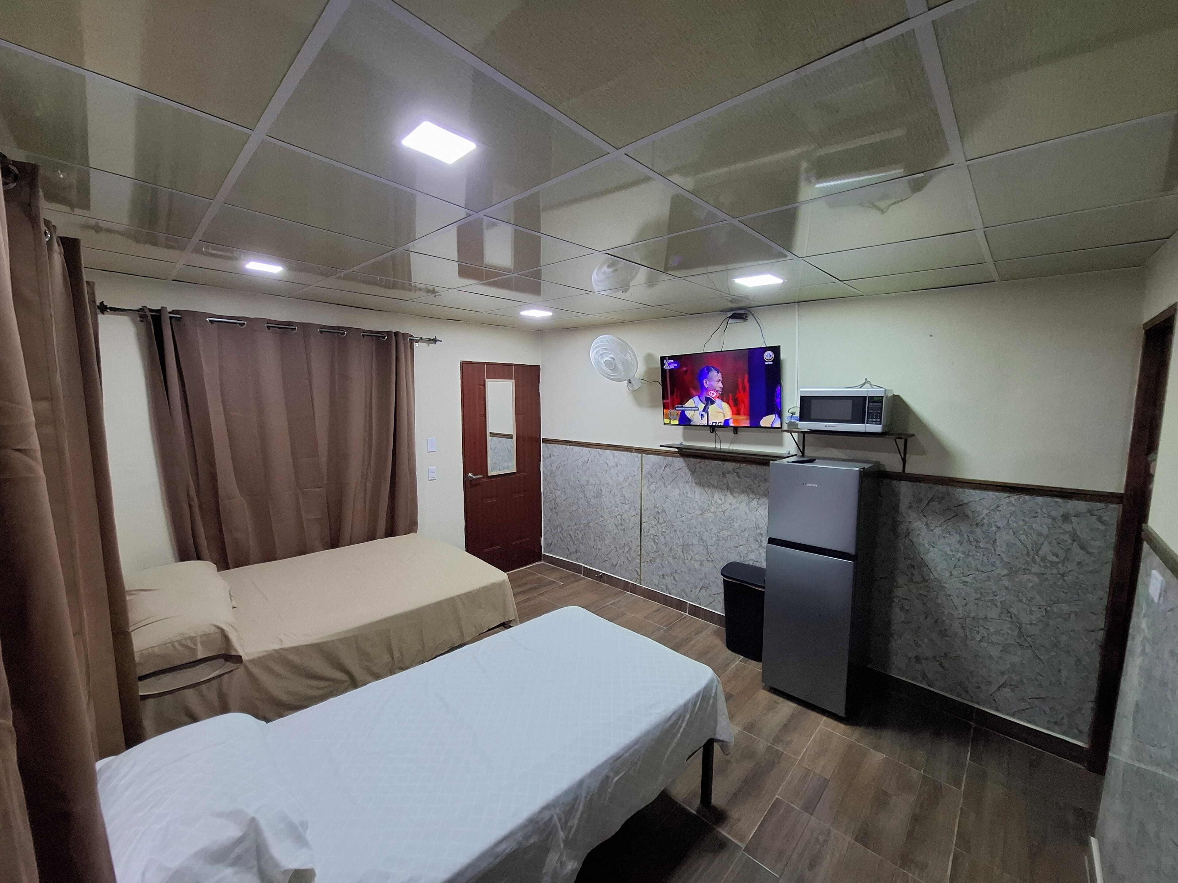 A compact hotel room with a bed, a small kitchenette, a TV, and a door.