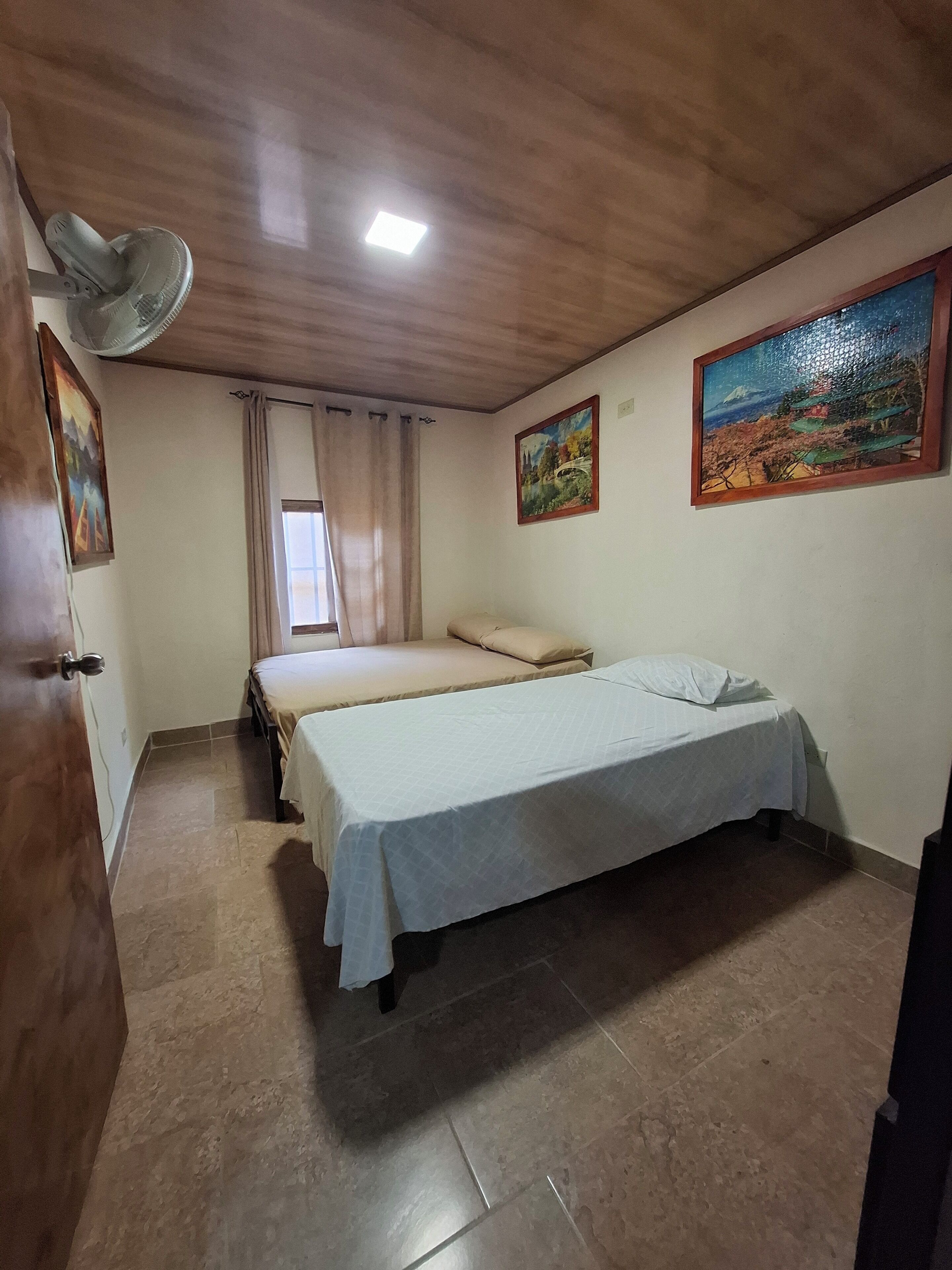 Una camera da letto con due letti, un ventilatore a soffitto, una finestra con tende e due quadri appesi al muro.
