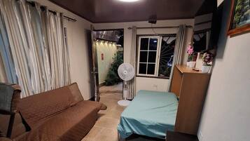Een kleine kamer met een bed, een slaapbank, een tafeltje en een ventilator.