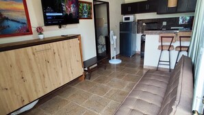 A spacious living area with a sofa, a TV mounted on the wall, a kitchen area with a refrigerator and microwave, and a wooden cabinet. - Aires frescos de Boquete (Alto Boquete)