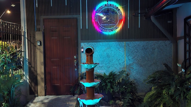 A fountain with multiple tiers in front of a building with a wooden door and a neon sign. - Aires frescos de Boquete (Alto Boquete)