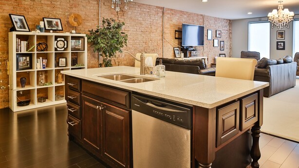 A kitchen with a central island, dark wooden cabinets, and a dishwasher. There is a brick wall and a television in the background. - 1 Bedroom Loft in Downtown Defiance! (Defiance)