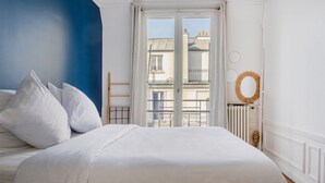 A bedroom with a bed, white bedding, a blue accent wall, a window with a view of buildings, and a radiator. - Courcelles Design Duplex (Paris)