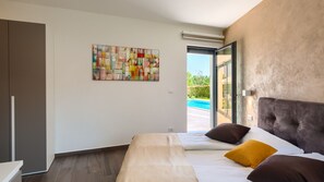 A modern bedroom with a large bed, a colorful abstract painting, and a view of a swimming pool through an open door. - Villa Nada by Villas Guide (Kringa)
