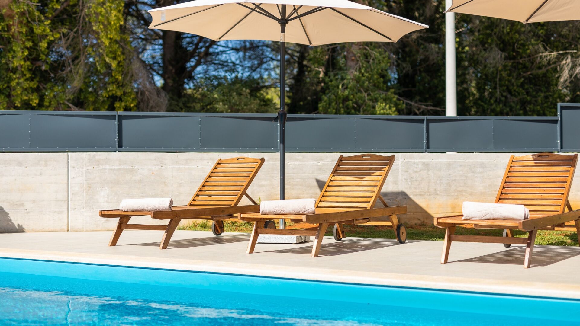 Ein Poolbereich mit drei Holzliegen und zwei beigen Sonnenschirmen.