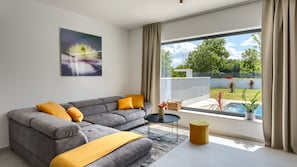 Un salon moderne avec un canapé gris, des coussins jaunes, une petite table ronde et une grande fenêtre donnant sur un jardin et une piscine.