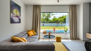Un salon moderne avec un canapé gris, des coussins jaunes, une table basse ronde et une vue sur une piscine et la verdure environnante.