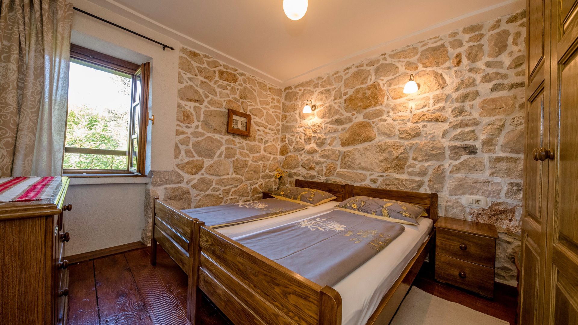 A bedroom with a stone wall, two beds, a nightstand, a wooden wardrobe, and a window with curtains.