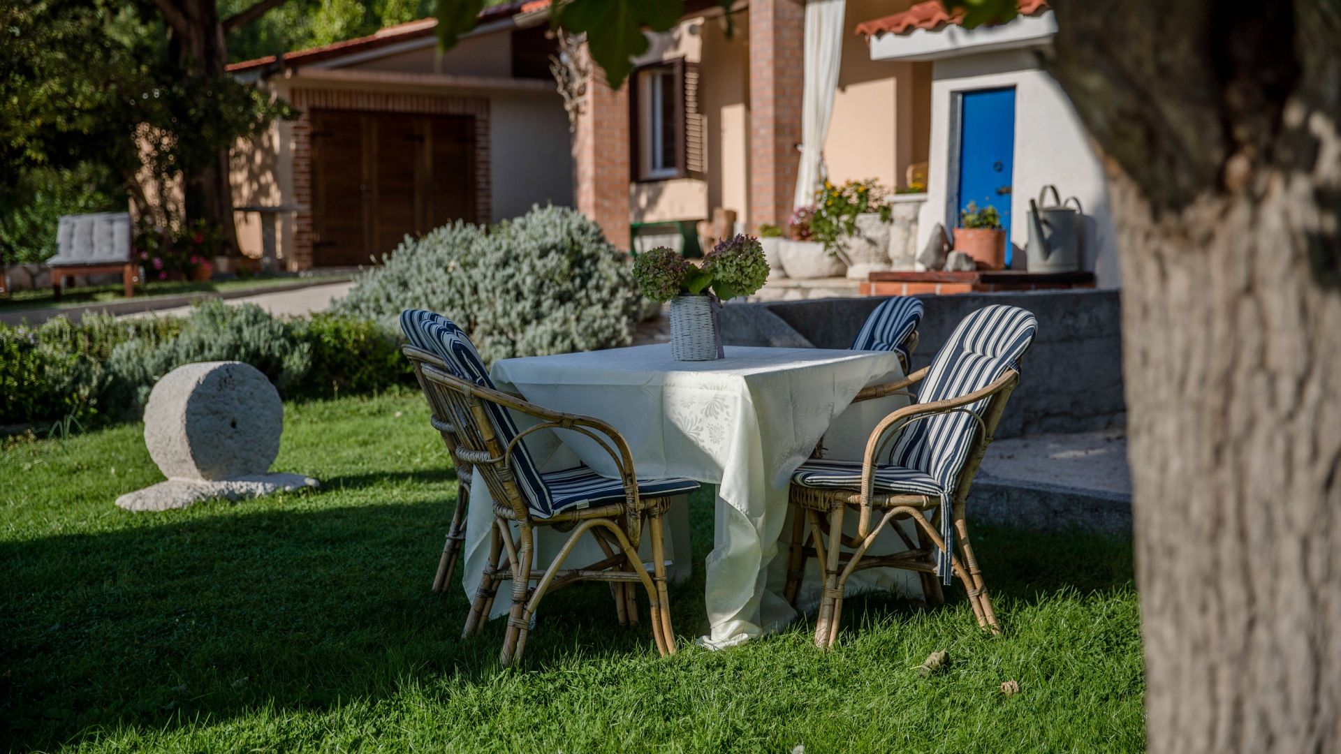 Un espace de restauration en plein air avec une table ronde et des chaises en osier, entouré d’une pelouse bien entretenue et d’une sculpture en pierre.