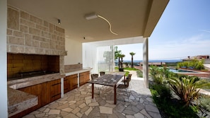 Un patio al aire libre con una chimenea de piedra, una mesa de madera y sillas, con vista a una piscina y un paisaje pintoresco.