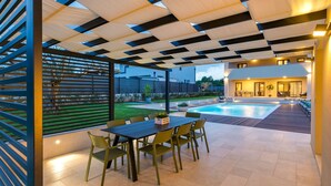 Ein überdachter Außenbereich mit Pool, einem Esstisch mit Stühlen und Blick auf ein modernes Haus.