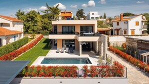 Een modern huis met twee verdiepingen, met een zwembad, omgeven door goed onderhouden tuinen en omliggende gebouwen.
