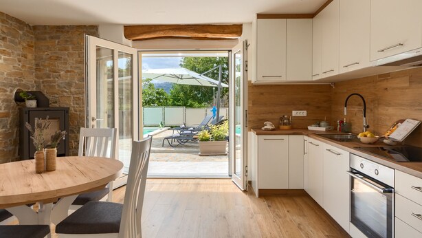 A modern kitchen with white cabinets, a wooden countertop, and a built-in oven. There is a dining area with a table and chairs, and a view of a pool and outdoor furniture through an open door. - Villa Casa Mavretici by Villas Guide (Livade)