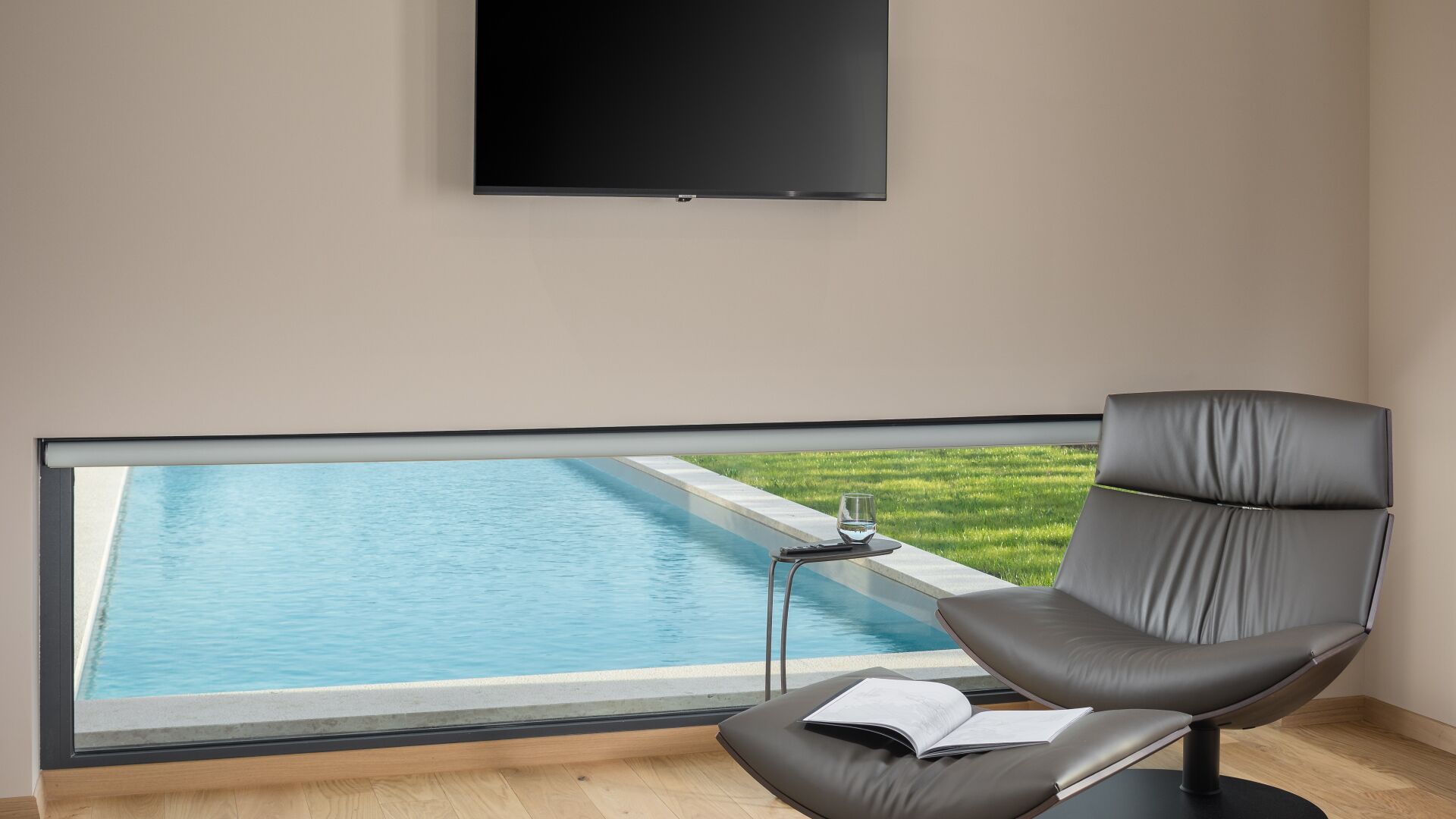 A room with a large window overlooking a swimming pool, a grey chair, and a flat-screen TV mounted on the wall.