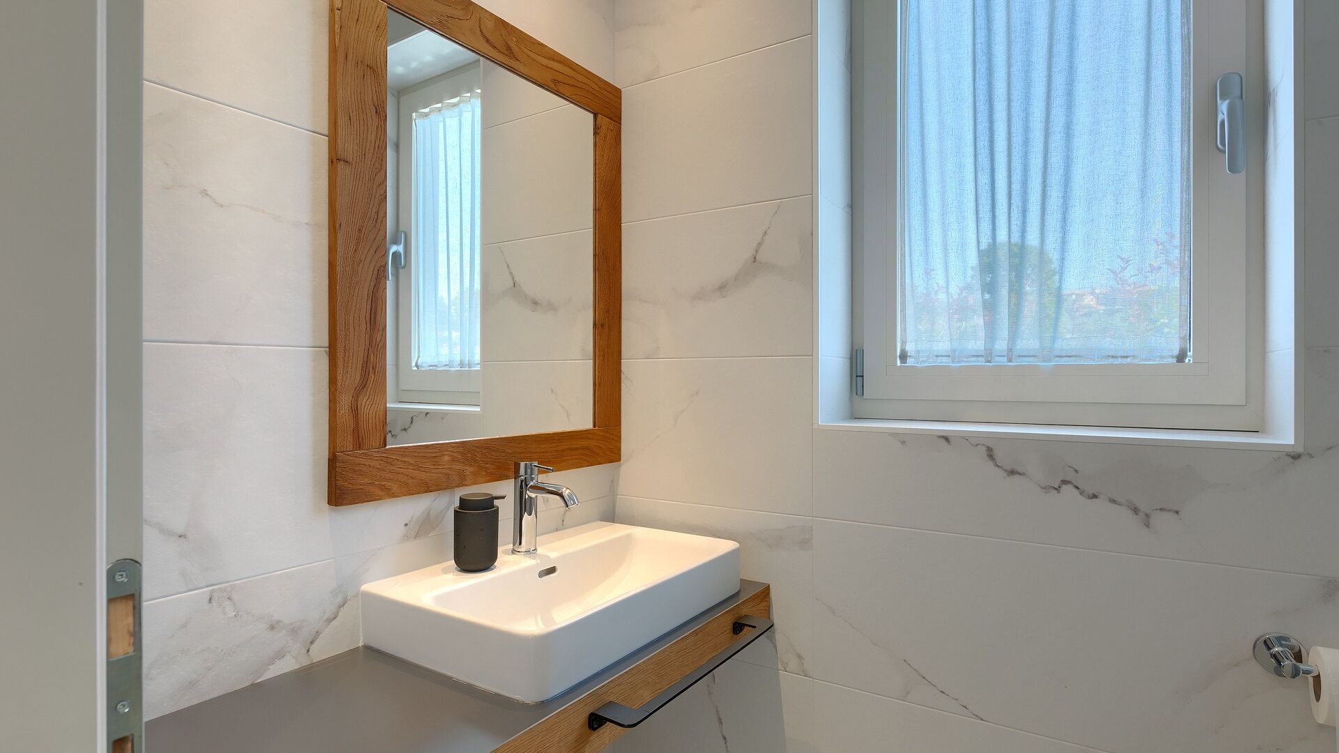 A modern bathroom with a white sink, wooden-framed mirror, and a window with sheer curtains.
