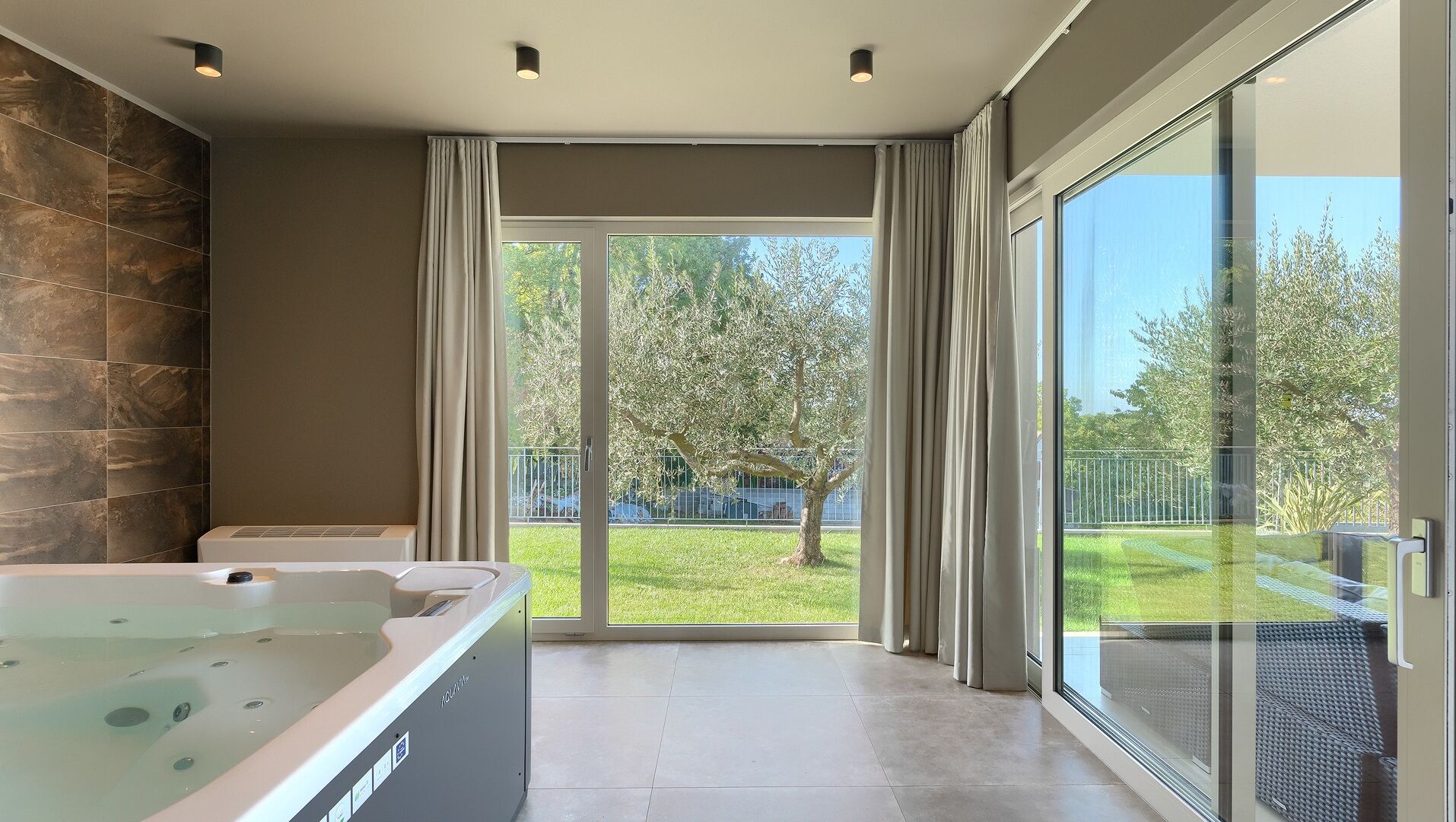 Ein modernes Badezimmer mit einer großen Badewanne, einer Steinwand und einer Glastür, die auf einen Balkon mit Blick auf einen Garten und Bäume führt.