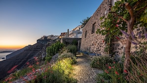 A stone pathway leading to a building with a stone wall and a view of the sea at sunset.