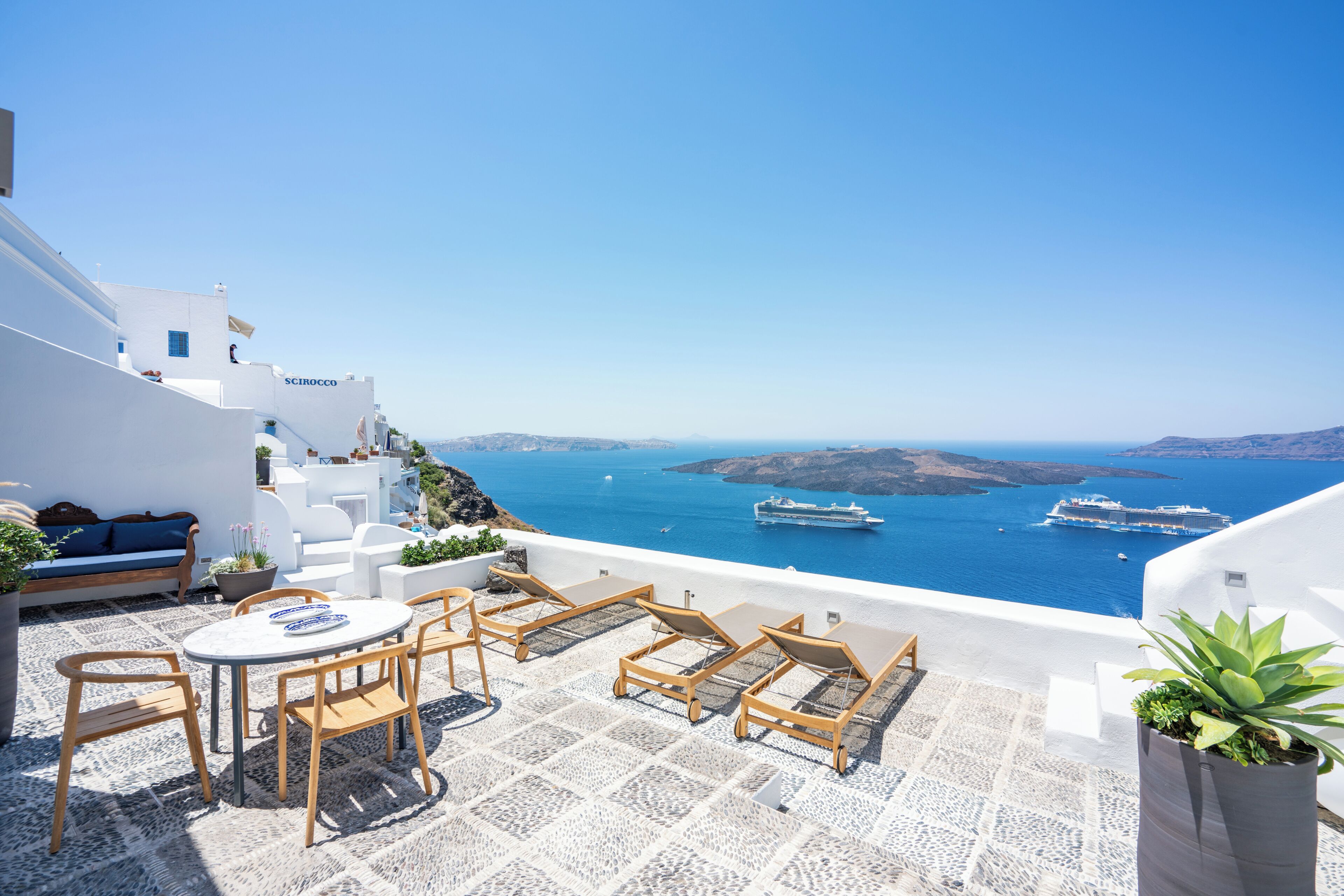 A terrace with a table and chairs, overlooking a sea with boats and a mountainous landscape.
