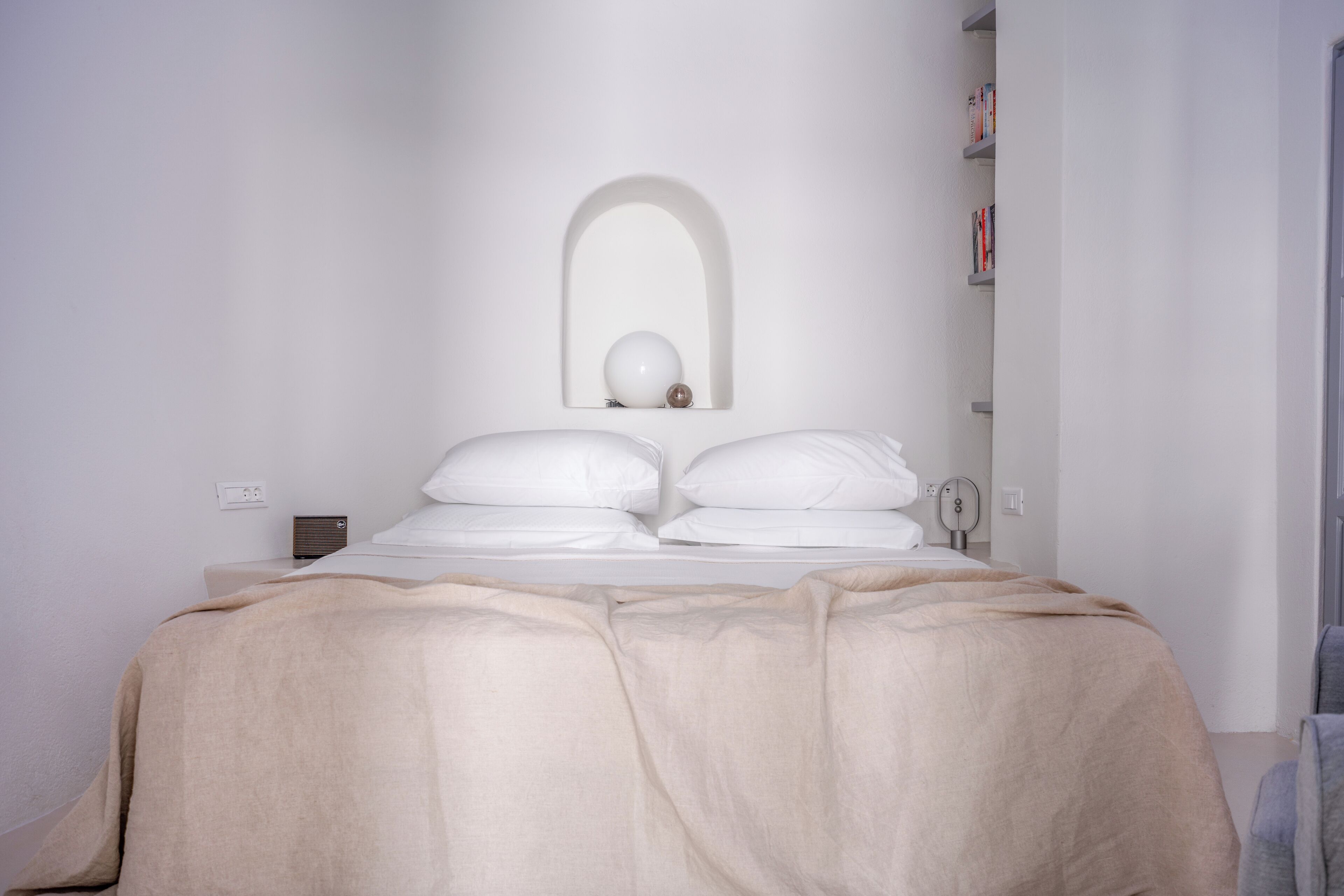 A minimalist bedroom with a bed, a bedside table, a mirror, and a wall-mounted shelf with books.