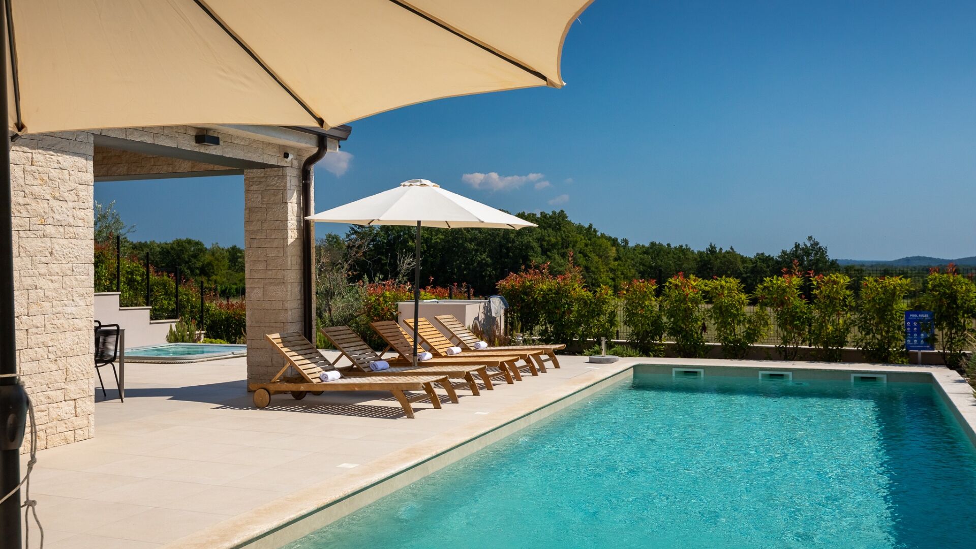 A poolside area with lounge chairs and a canopy, surrounded by greenery and a clear sky.
