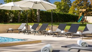 A poolside area with lounge chairs, umbrellas, and a children's play structure in the background. - Villa Corticum Otium by Villas Guide (Kringa)