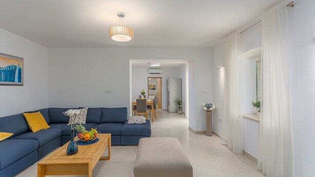 A modern living room with a blue sofa, a wooden coffee table with fruit, a small table with a vase, a dining area in the background, and a hallway leading to another room. - Villa Corticum Otium by Villas Guide (Kringa)
