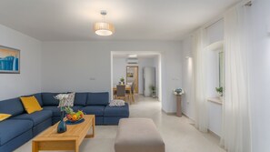 A modern living room with a blue sofa, a wooden coffee table with fruit, a small table with a vase, a dining area in the background, and a hallway leading to another room. - Villa Corticum Otium by Villas Guide (Kringa)