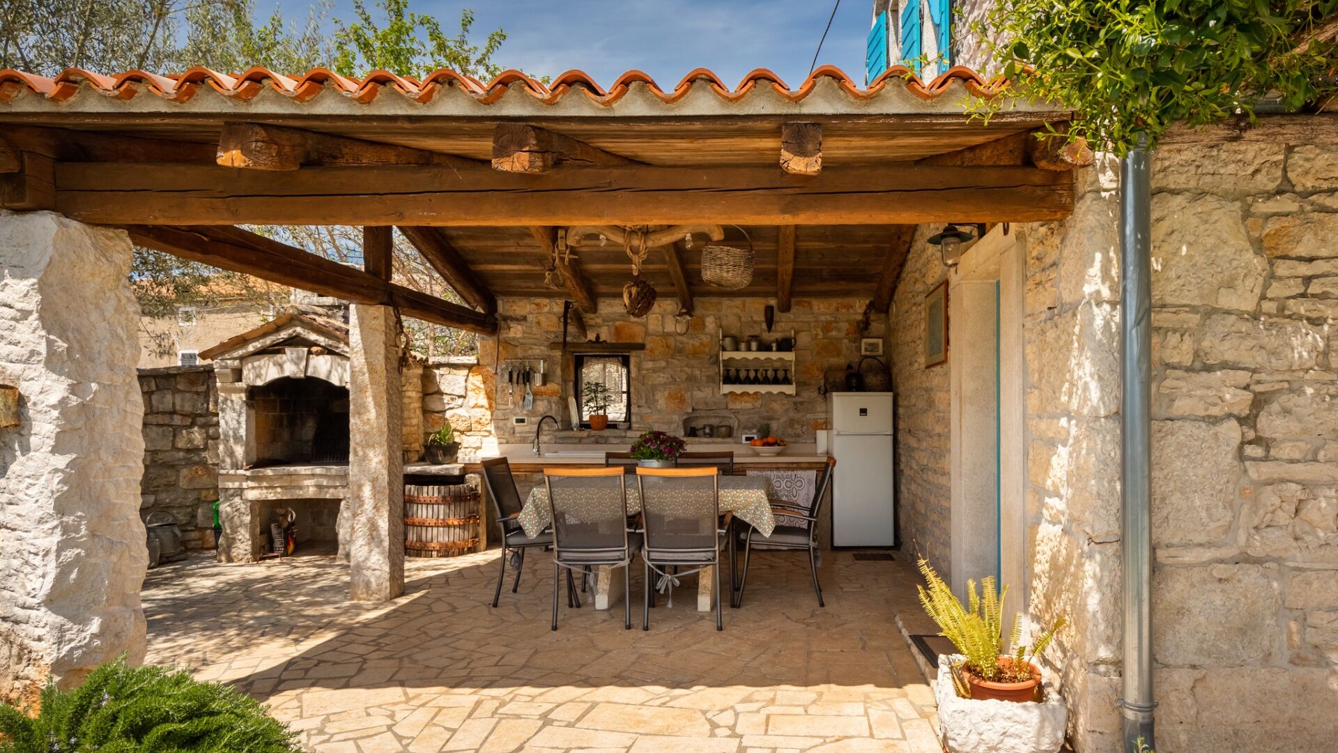 A rustic outdoor kitchen with stone walls, a wooden pergola, and a stone fireplace.