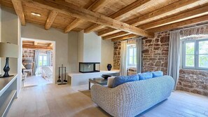 Quarto com vigas de madeira aparentes, parede de pedra, sofá, televisão e janela com cortinas.