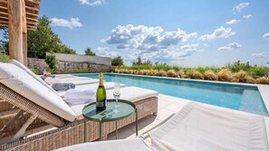 A poolside area with a wicker lounge chair, a glass table with a bottle and glass, and a view of the pool and sky.
