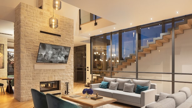 A modern living room with a stone fireplace, a large flat-screen TV, a grey sofa, and a staircase with glass railings.