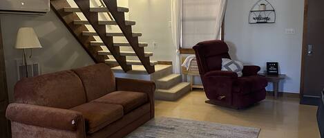 Un salon avec un canapé marron, un fauteuil rouge, un escalier en bois et une pancarte « Bienvenue » accrochée au mur.