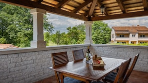 Alpendre coberto com mesa posta para refeições, paredes de pedra e vista para um edifício com telhado em cerâmica.