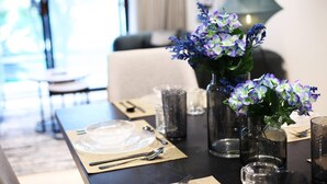 Une table à manger dressée avec de la vaisselle transparente, un vase rempli de fleurs violettes et un intérieur moderne avec un canapé et une lampe en arrière-plan.