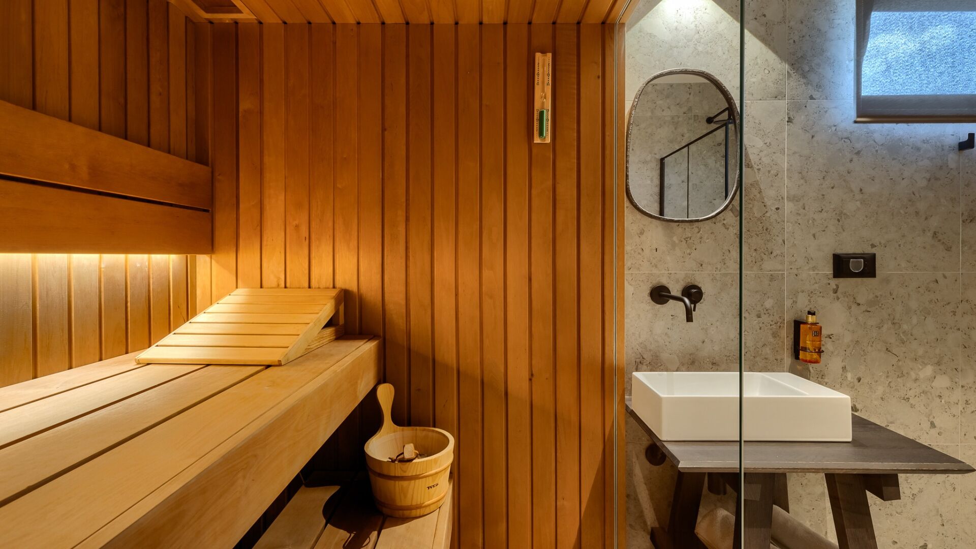 A wooden sauna with benches and a wooden bucket, a glass-enclosed shower area with a white sink, and a small window.