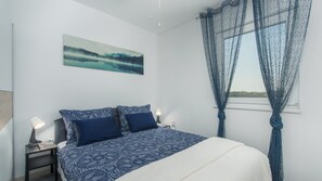 Een slaapkamer met een bed, twee nachtkastjes, een lamp, een schilderij en een raam met dunne gordijnen.
