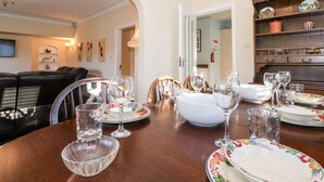 Un coin repas avec une table en bois dressée pour un repas, entourée de chaises. On aperçoit une bibliothèque avec des objets décoratifs et un téléviseur au fond.