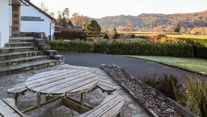 Une terrasse en pierre avec une table et des bancs en bois, un bâtiment blanc avec un escalier, et une vue sur un paysage verdoyant avec des collines en arrière-plan.