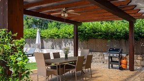 An outdoor patio with a table and chairs, a ceiling fan, a grill, and a stone wall.