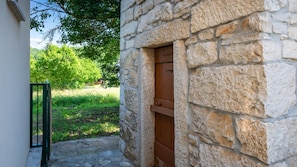 A stone building with a wooden door, a metal gate, and a grassy area with trees in the background.