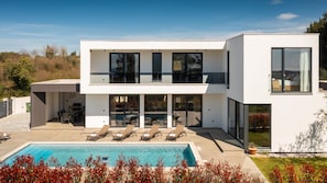 Ein modernes, zweistöckiges Haus mit Swimmingpool, Sitzgelegenheit im Freien und großen Fenstern.