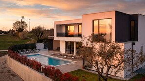 Ein modernes, zweistöckiges Haus mit Swimmingpool, Sitzgelegenheit im Freien und Blick auf den Sonnenuntergang.