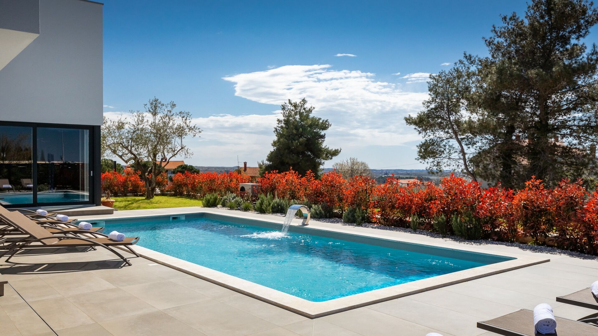 A rectangular swimming pool with a water feature, surrounded by lounge chairs and a garden with red foliage.