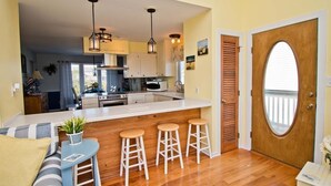 A modern kitchen with a central island, wooden flooring, and a dining area with a sofa and bar stools. - Dark and Stormy, Almost Oceanfront House in Emerald Isle, NC! (Emerald Isle)
