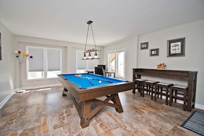Una mesa de billar con superficie de fieltro azul, base de madera y una lámpara colgante. Cuatro taburetes de madera están alineados frente a un banco de madera oscura.