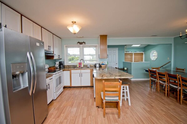 A modern kitchen with stainless steel appliances, a central island, and a dining area with wooden chairs. - Decks Ahoy, Oceanfront House in Emerald Isle, NC! (Emerald Isle)