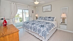 A bedroom with a bed, a ceiling fan, a window with curtains, a bedside table with a lamp, and a view of the outside. - Dock Holiday, Oceanview House in Atlantic Beach, NC! (Atlantic Beach)