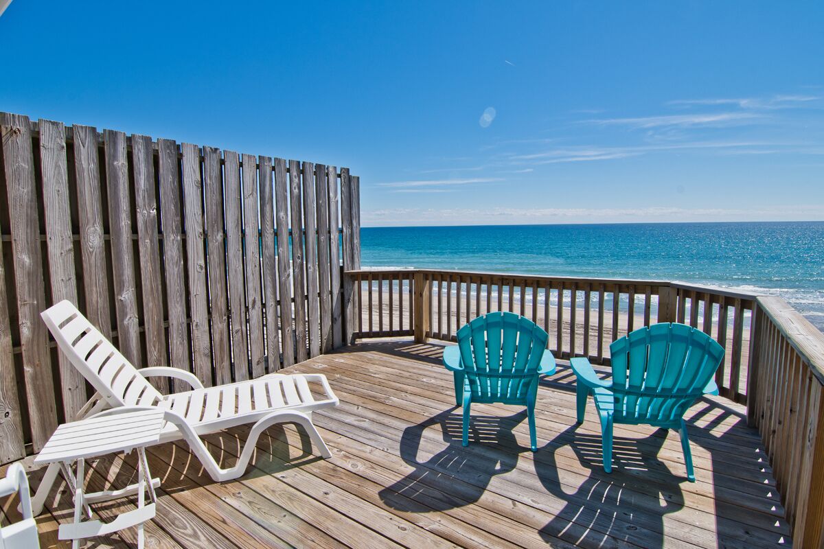 A wooden deck with two green Adirondack chairs and a white lounge chair, overlooking the ocean.